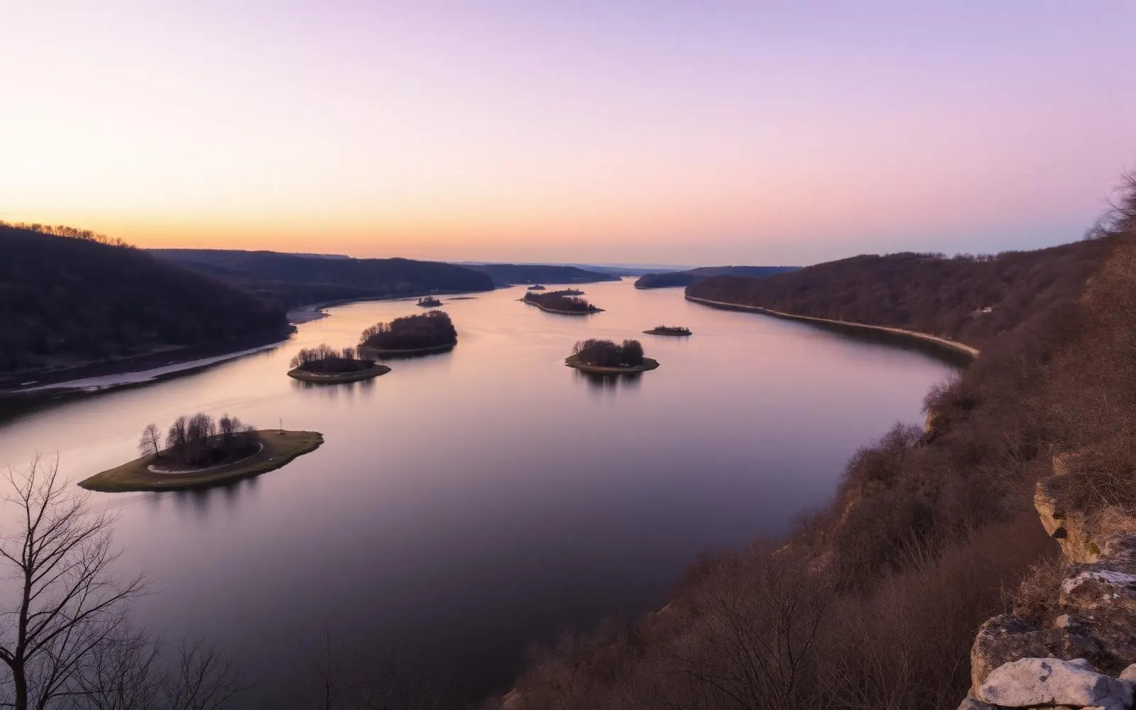 The wide Susquehanna River at dusk seen from a wooded bluff, with low islands and a soft violet and gold sky