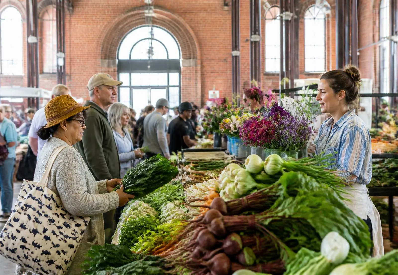 Shoppers and a vendor talking across a produce stall inside a brick market house